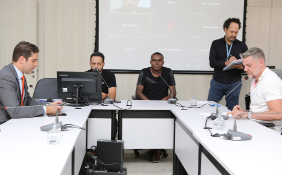 Quatro vereadores sentados à mesa, em reunião presencial, acompanhados de Assessor da Câmara, de pé. 