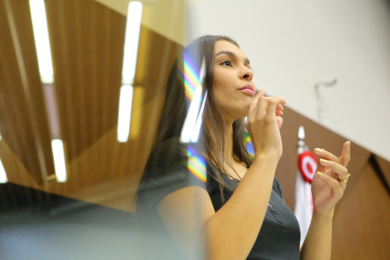 Mulher caucasiana utiliza língua de sinais. Ela veste blusa presta e tem cos cabelos longos, pretos e lisos