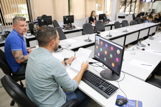 Dois vereadores sentados à mesa em reunião presencial, um deles tem tela de computador à frente na qual outros vereadores participam de maneira remota.