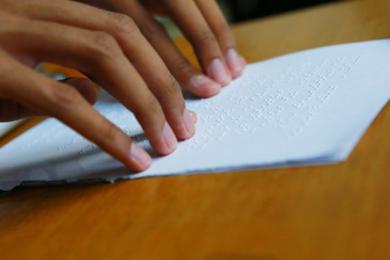 As mãos de uma pessoa percorrem um papel branco escrito em braile