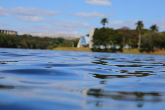 Imagem do espelho d'água da Lagoa, em primeiro plano. Ao fundo a imagem desfocada da igrejinha da Pampulha 