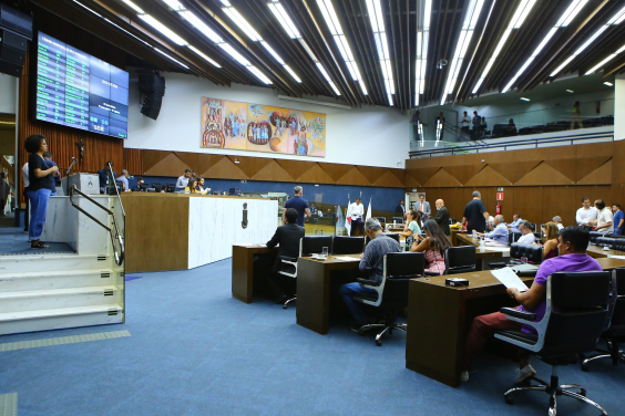 Imagem lateral do Plenário Amynthas de Barros. Estão enquadrados a mesa diretora e as cadeiras dos parlamentares. Ao fundo e no alto, o painel de Yara Tupinambá
