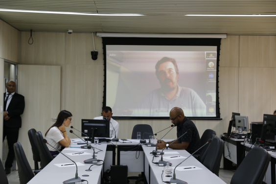 Imagem dos parlaementares sentados em uma mesa em formato de U. Ao fundo e no alto, um telão exige a imagem da testemunha