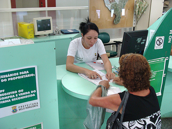 Imagem de atendimento na Central de Atendimento Barreiro. A atendente uniformizada com uma blusa branca e a logo da PBH atende uma mulher que se encontra de blusa preta. Ambas estão sentadas de frente uma para a outra