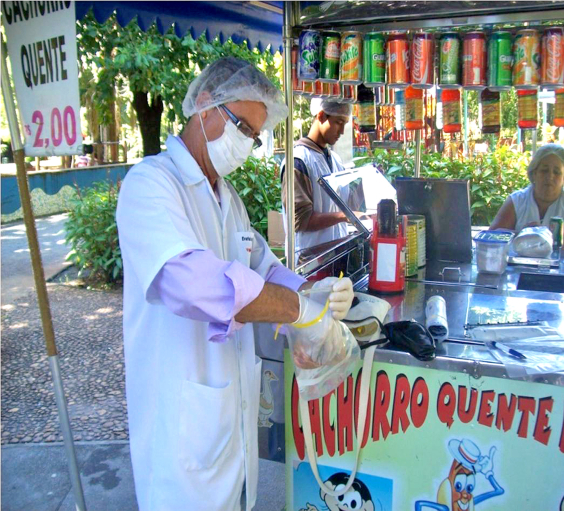 Imagem de vendedor ambulante em uma barraca de bebidas e cachorro quente. Ele usa jaleco, touca e luvas brancas