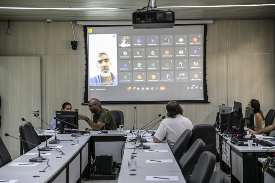 Três vereadores participam de reunião, sentados à mesa. No telão, é exibida reunião vistual com mais de 20 participantes, entre eles o secretário municipal de Planejamento, Orçamento e Gestão, André Reis.