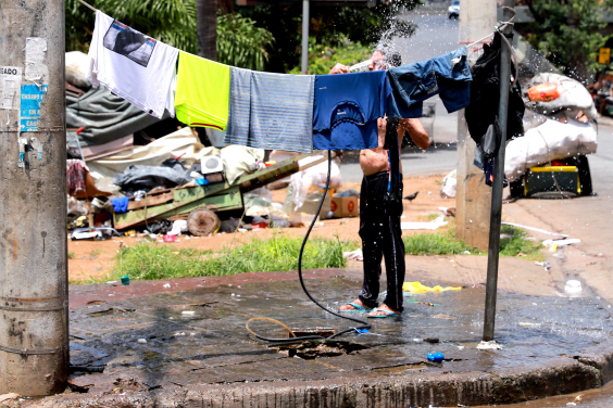 Imagem de uma pessoa tomando banho de mangueira. ela está atrás de um varal cheiro de roupas que secam ao ar livre, em plena praça