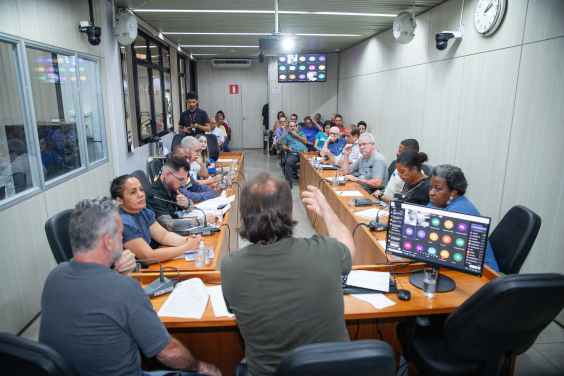 Dois vereadores e uma deputada estadual reunidos à mesa com dez cidadãos, com maid e dez pessoas sentadas em cadeiras ao fundo da sala.