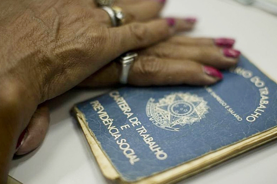 Mãos femininas sobre uma carteira de trabalho bastante usada, por cima de uma mesa.