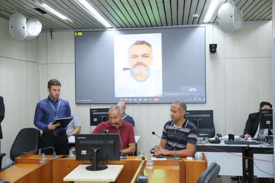 Dois parlamentares sentados à mesa, em reunião. Ao fundo, parlamentar que participa da reunião de maneira remota tem sua imagem exibida em telão.