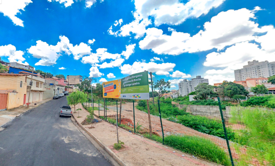 Rua urbanizada com placa de obra no córrego Lareira, à direita.