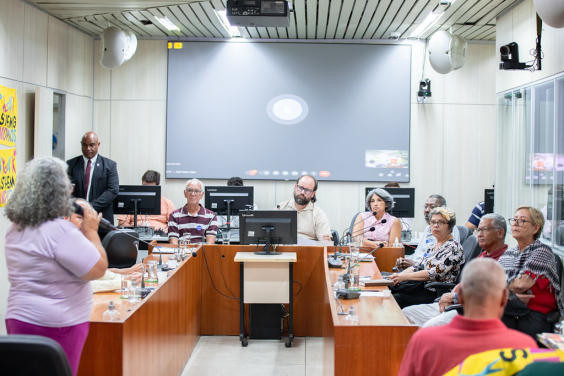 Quatro homens e três mulheres reunidos, sentados à mesa, assistem a mulher que fala, de pé.