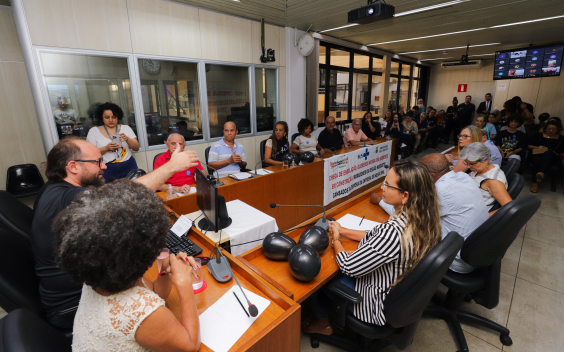 Dose pessoas reunidas, sentadas à mesa, e mais de vinte sentadas ao fundo.
