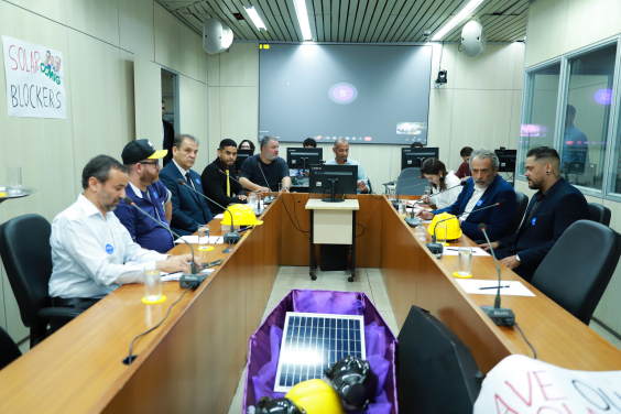 Nove pessoas reunidas, sentadas à mesa. No centro, um caixão uma célula de produção de energia solar acompanhada por capacetes de construção.