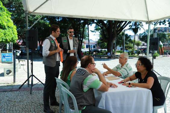 Um homem e uma mulher se consultam com profissionais da Ouvidoria Itinerante sentados à mesa, em praça, durante o dia, acompanhados pelo ouvidor e outro servidor da Ouvidoria. 