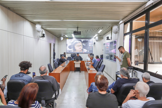 Quatro homens e uma mulher reunidos, sentados à mesa; em telão do fundo, mulher participa de reunião de forma remota. À frente, mais de oito pessoas acompanham a reunião, sentados. 