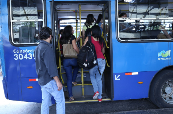 A foto mostra a lateral da parte de traseira de um ônibus azul. A porta está aberta, com pessoas dentro e outras preste a entrar.