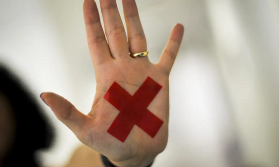 foto mostra uma mão aberta, com a palma virada para frente, uma aliança no dedo anelar e um "X" vermelho colado na palma.