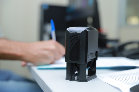 A foto mostra um carimbo em cima de uma mesa. Atrás dele, uma mão escreve à caneta.