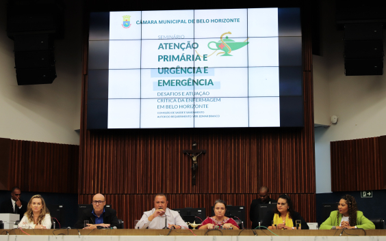 Membros da mesa de debate do seminário de enfermagem estão assentados no Plenário Amintas de Barros; acima deles está um telão com um slide