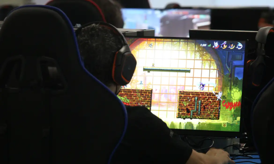 Na foto, no canto esquerdo, observa-se parcialmente a parte de trás de uma cadeira e um jovem sentado utilizando um fone de ouvido. Na sua frente a tela de um computador mostra um jogo.