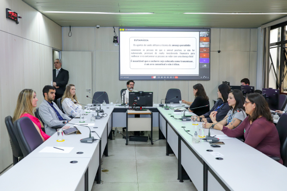 Osvaldo Lopes e convidados da audiência pública assentados no Plenário Helvécio Arantes, tela atrás deles mostra apresentação sendo realizada