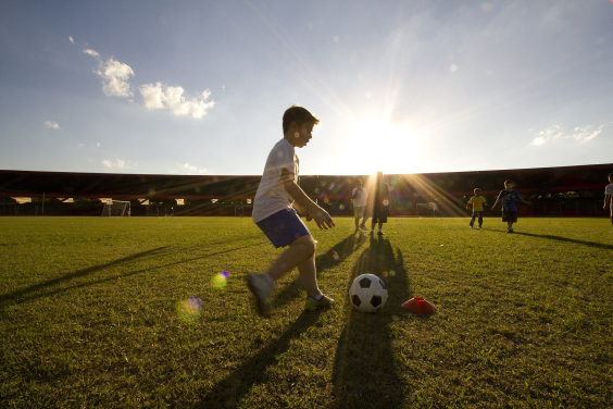 Menino chuta bola em campo de futebol próximo