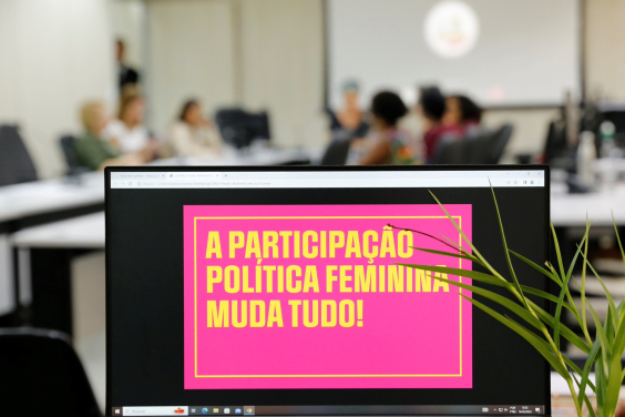 Imagem mostra frase "a participação feminina muda tudo" exibida em computador com audiência pública na CMBH acontecendo ao fundo