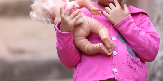 Imagem de uma criança pequena com dedo na boca, usando blusa rosa e carregando uma boneca. 