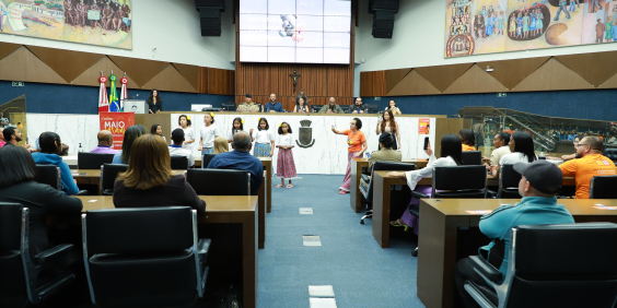 Acompanhada por adulta, cinco crianças de saia de chita e flor no cabelo se apresentam no Plenário Amintas de Barros.