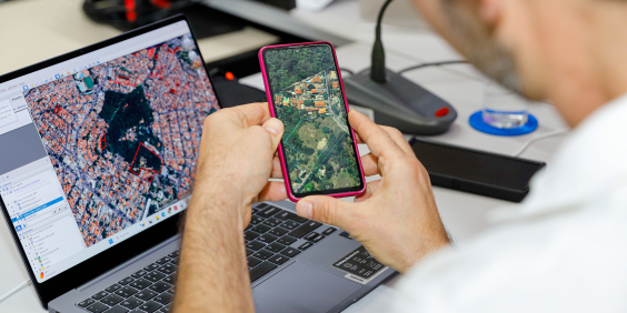 homem com o celular na mãe e o computador à frente, ambos com imagem geográfica de satélite