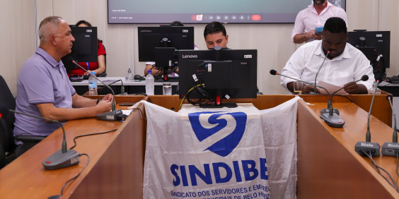 Vereadores em reunião no Plenário Camil Caram; bandeira do Sindibel está erguida na frente da mesa do presidente da comissão