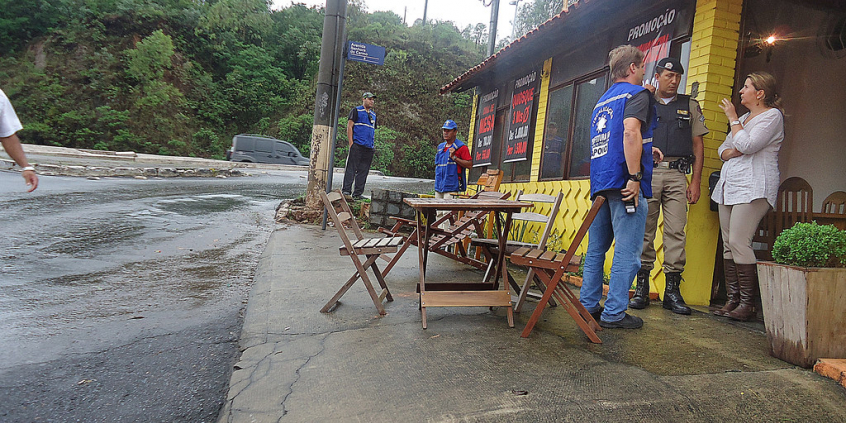 A imagem mostra um fiscal da Prefeitura em frente a um bar com mesas na calçada 