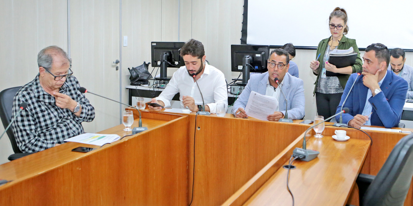 Integrantes da Comissão estão sentados à Mesa apreciando a pauta da reunião. Atrás, há uma servidora de pé, assessorando a reunião