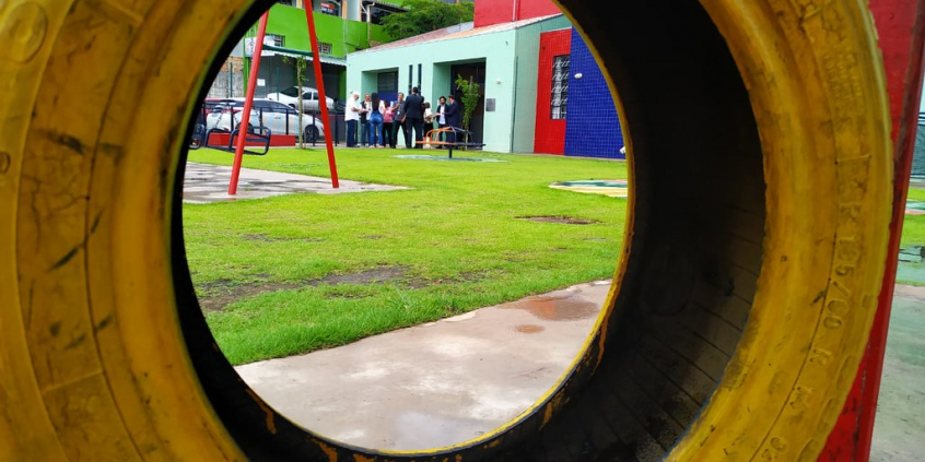 em destaque, gangorra em pneu amarelo. Ao fundo, pátio de recreação colorido, gramado e comitiva parlamentar de pé