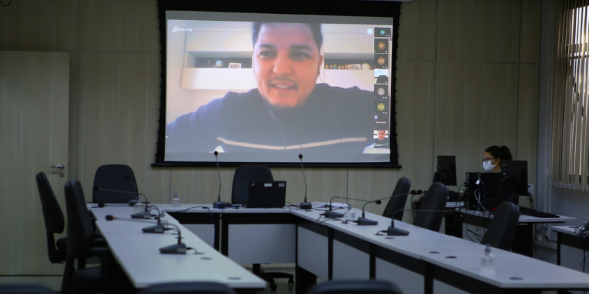 A imagem mostra a mesa do plenário vazia, com a equipe de assessoria ao lado, e um telão com as imagens dos vereadores em participação remota