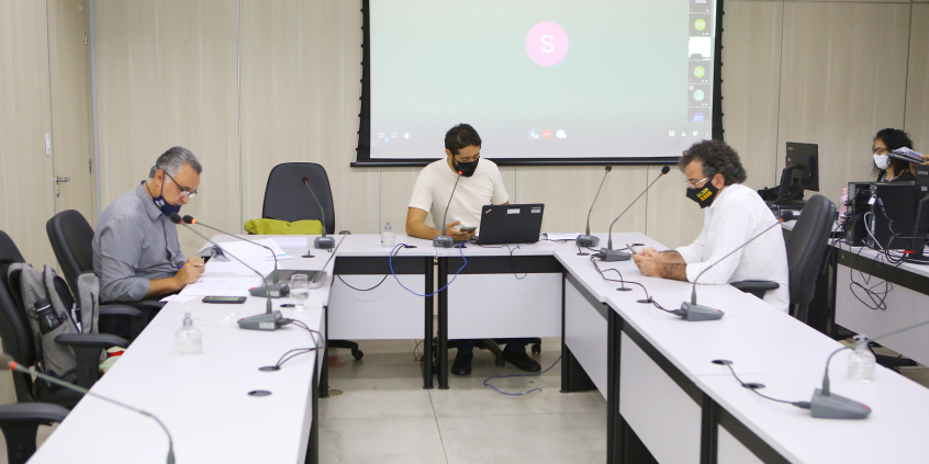 Três vereadores sentados à mesa e telão ao fundo em videoconferência com convidados