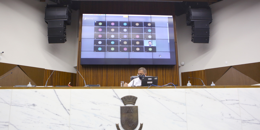 Parlamentar compõe mesa de reunião. Telão ao fundo em videoconferência com os demais membros e convidados.