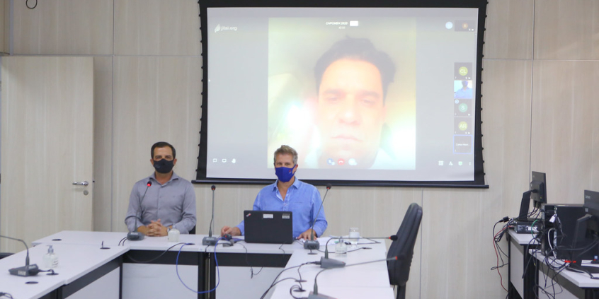 Parlamentares compõem mesa de reunião. Telão ao fundo em videoconferência com os demais membros