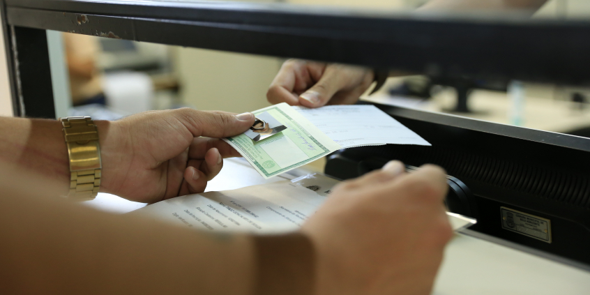 Detalhe das mãos do usuário e da funcionária do Posto de Identificação no momento do atendimento no guichê. Usuário recebe o novo documento de identidade