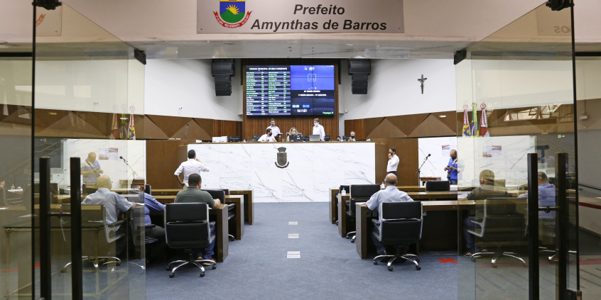 vista geral do Plenário Amynthas de Barros_vista a partir da porta de entrada. Porta de vidro aberta, em primeiro plano. Ao fundo, vereadores ocupam os seus lugares. No alto, placa indica o nome do Plenário