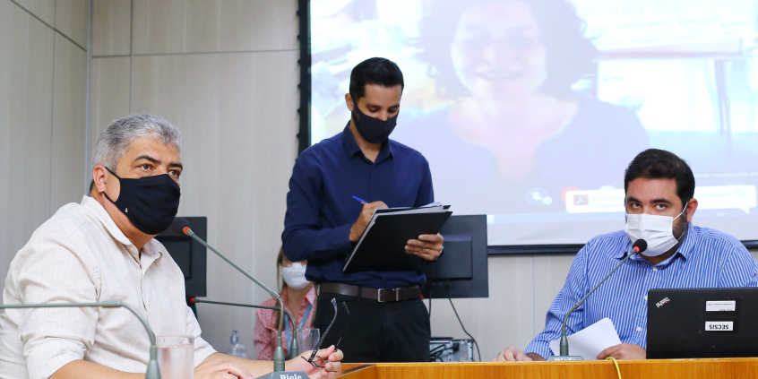 Os vereadores professor Cludiney Dulim e Bruno Miranda sentados à mesa, de máscara, próximos a um assessor da Câmara, de pé, também de máscara. Ao fundo, a imagem vereadora Sônia Lansky da Coletiva em um telão.