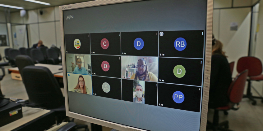 Monitor de computador exibe convidados em videoconferência. Ao fundo, cadeiras do Plenário Helvécio Arantes, vazias