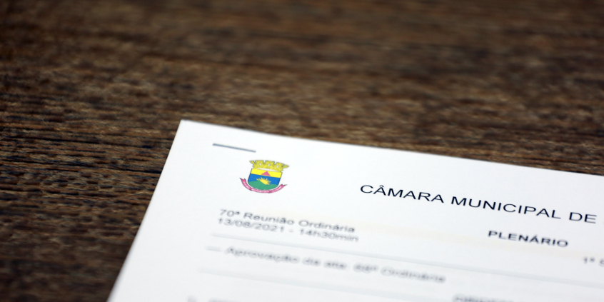 Detalhe da pauta da reunião impressa, sobre a mesa de madeira. Cabeçalho exibe brasão de belo horizonte e assinatura da Câmara Municipal