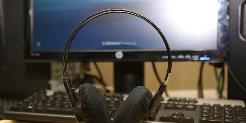 Fone de ouvido em pé, em cima da mesa. ao fundo, tela de computador