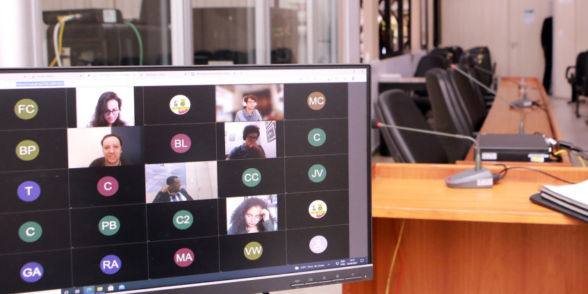 tela exibe parlamentares e convidados em reunião por videoconferencia. ao fundo, plenário vazio