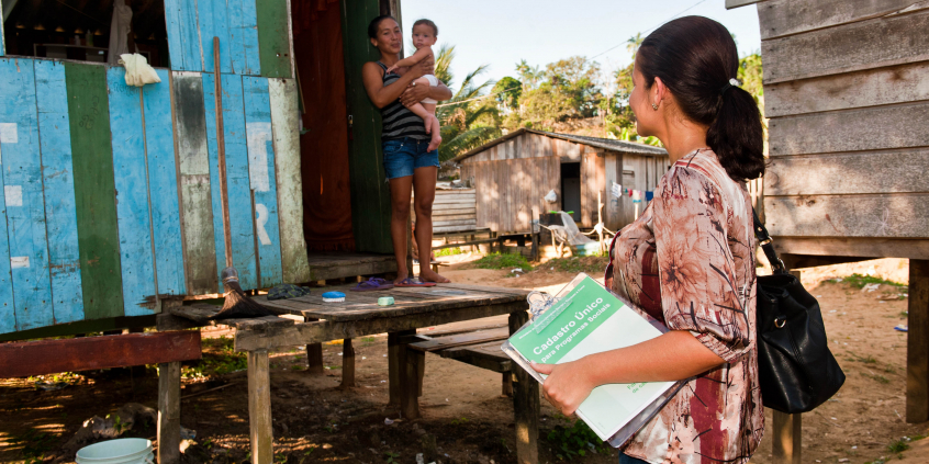 Profissional de assistência social visita área carente da cidade com caderno de anotações do Cadastro único, durante o dia.