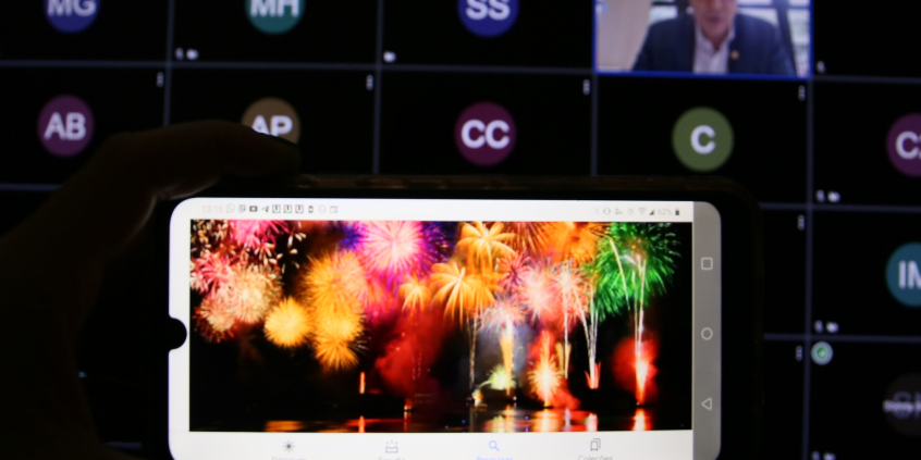 Imagem das luzes dos fogos de artifícios na tela do celular. Ao fundo a tela do comutador com a imagem dos participantes da audiência