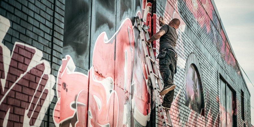 Homem em cima de escada, pinta muro urbano com desenhjos coloridos, durante o dia