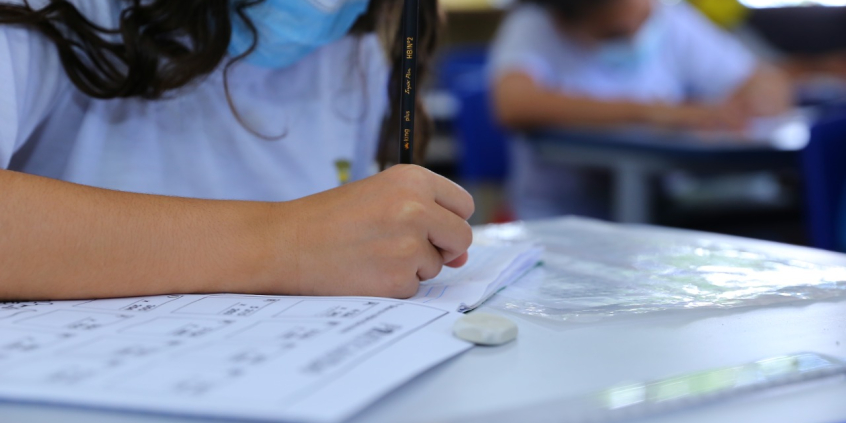 Menina sentada, inclinada sobre carteira escolar, com máscara no rosto, preenche um caderno educativo com lápis. Ao fundo, outro estudante realiza a mesma atividade,